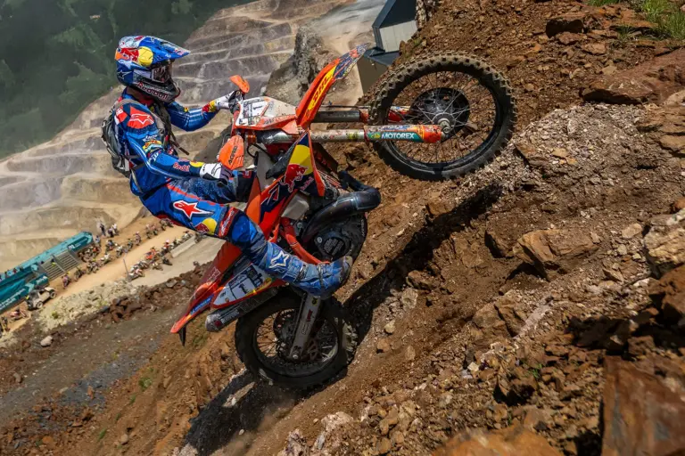 Erzbergrodeo: Fünfter Streich zum Jubiläum Ein Enduro-Motorradfahrer in blau-roter Schutzausrüstung kämpft sich eine extrem steile, felsige Abraumhalde hinauf. Das Vorderrad des orangefarbenen Motorrads hebt sich über eine Geländekante, während Geröll unter den Reifen wegrutscht. Im Hintergrund sind die terrassenförmigen Stufen eines großen Tagebaus sowie weitere Motorräder und Zuschauer zu erkennen. Die Aufnahme vermittelt Dynamik, Kraft und die Herausforderung des anspruchsvollen Offroad-Wettbewerbs.