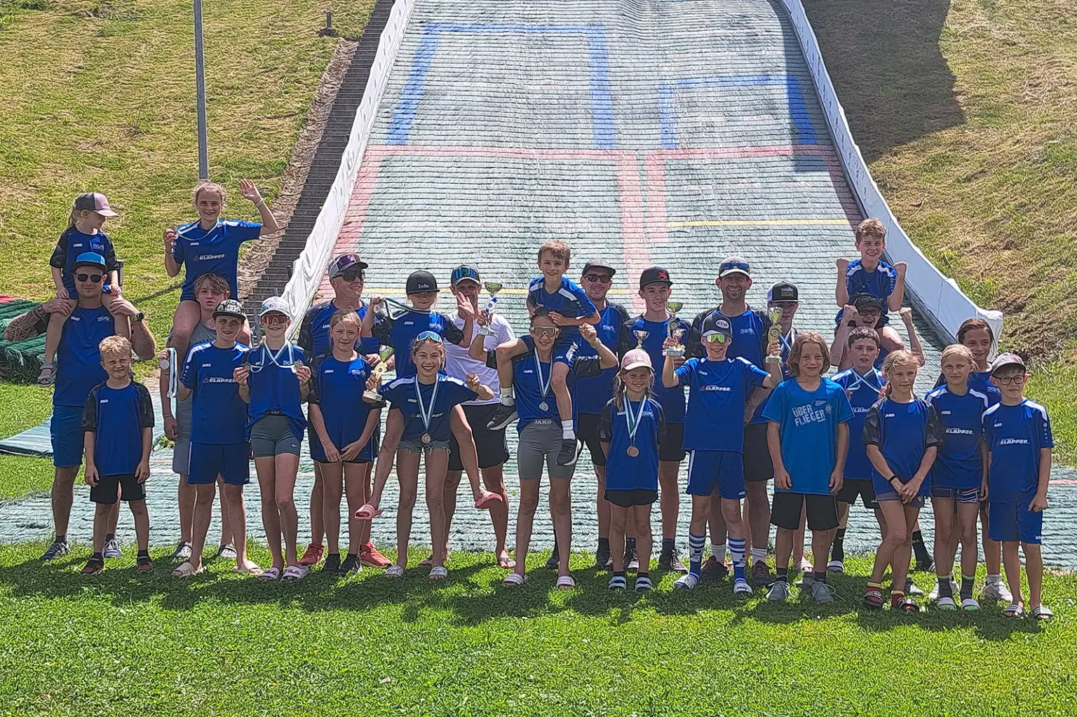 Gruppe von Kindern und Erwachsenen in blauen Vereinsdressen vor einer Skisprungschanze. Mehrere Kinder halten Pokale und Medaillen, einige jubeln in die Kamera. Im Hintergrund ist die Schanze zu sehen.