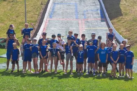Gruppe von Kindern und Erwachsenen in blauen Vereinsdressen vor einer Skisprungschanze. Mehrere Kinder halten Pokale und Medaillen, einige jubeln in die Kamera. Im Hintergrund ist die Schanze zu sehen.