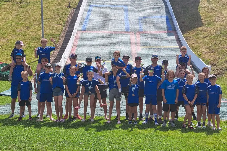 Sechs Tagessiege für den SC Erzbergland Gruppe von Kindern und Erwachsenen in blauen Vereinsdressen vor einer Skisprungschanze. Mehrere Kinder halten Pokale und Medaillen, einige jubeln in die Kamera. Im Hintergrund ist die Schanze zu sehen.