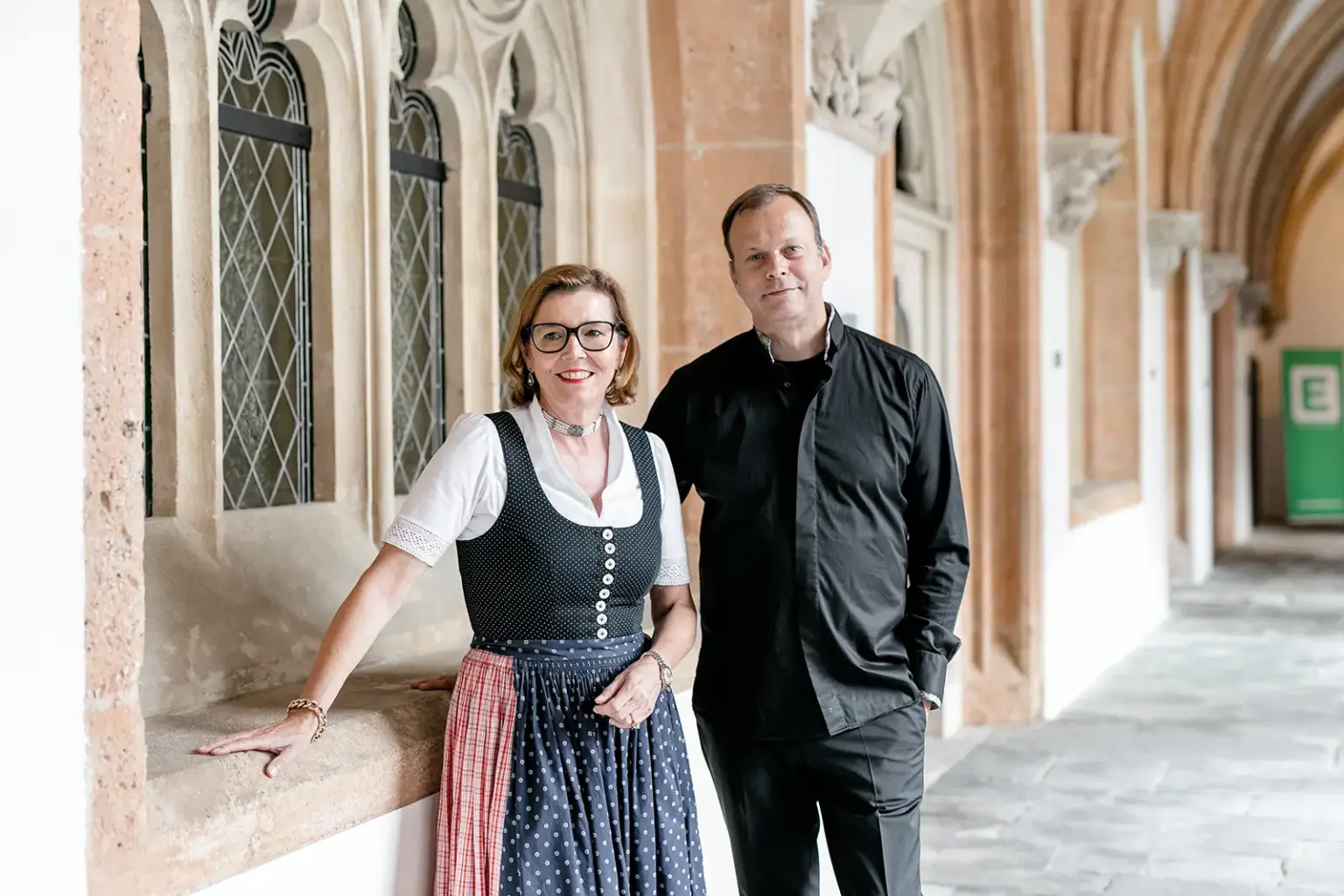 Eine Frau in Tracht steht neben einem Mann im schwarzen Gewand in einem Kreuzgang eines Stiftes.