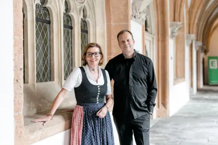 Eine Frau in Tracht steht neben einem Mann im schwarzen Gewand in einem Kreuzgang eines Stiftes.