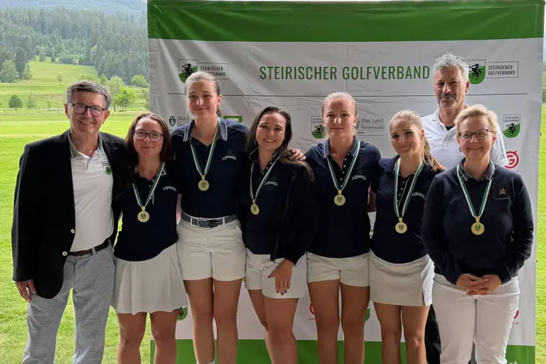 Eine Golfmannschaft mit Medaillen steht vor einer Sponsorenwand des Steirischen Golfverbands für ein Gruppenfoto. Die Spielerinnen und Betreuer posieren gemeinsam, im Hintergrund sind der Golfplatz und eine bewaldete Hügellandschaft zu sehen.