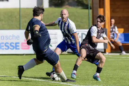 Die Relegationsspiele sind fixiert Drei Fußballspieler kämpfen auf dem Rasen um den Ball. Während ein Spieler in blau-weiß gestreiftem Trikot nach vorne drängt, versuchen zwei Gegenspieler in dunklen Dressen den Angriff zu stoppen.