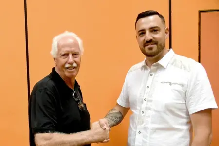 Zwei Männer stehen vor einer orangefarbenen Wand, geben sich die Hand und blicken in die Kamera.
