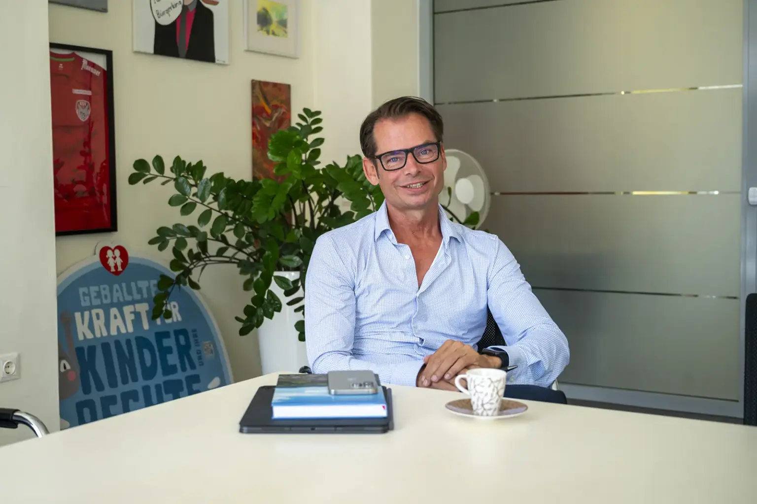 „Wir sind besser, als wir jahrelang geglaubt haben“ Ein Mann mit Brille sitzt an einem Besprechungstisch in einem Büro und lächelt in die Kamera. Auf dem Tisch liegen ein Laptop, Bücher und eine Kaffeetasse, im Hintergrund sind Pflanzen, Bilder und eine Glastür zu sehen.