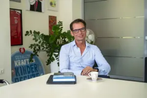 „Wir sind besser, als wir jahrelang geglaubt haben“ Ein Mann mit Brille sitzt an einem Besprechungstisch in einem Büro und lächelt in die Kamera. Auf dem Tisch liegen ein Laptop, Bücher und eine Kaffeetasse, im Hintergrund sind Pflanzen, Bilder und eine Glastür zu sehen.