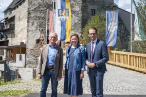 Drei Personen stehen vor einer historischen Burganlage und posieren für ein Foto. Im Hintergrund sind Fahnen, eine gepflasterte Zufahrt und die alten Steinmauern der Burg zu sehen.