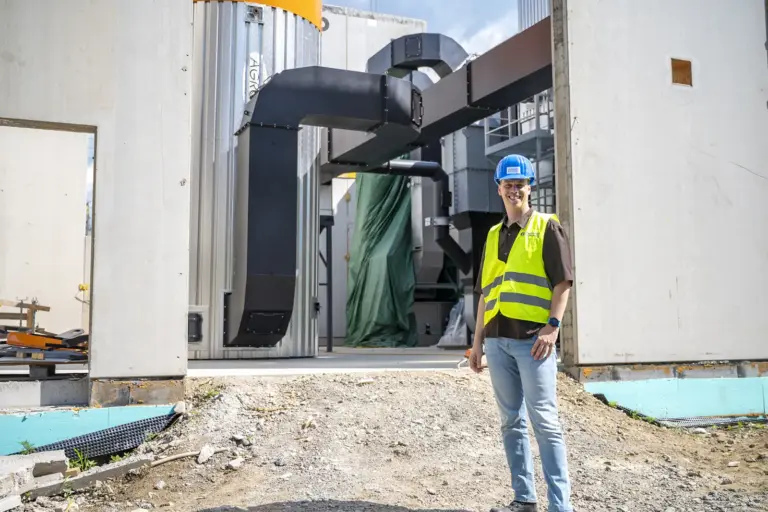 Kapfenberger Stadtwärme-Ausbau läuft nach Plan Ein Mann mit blauem Schutzhelm und gelber Warnweste steht vor einer Industrieanlage im Rohbau. Hinter ihm sind große Metallrohre, technische Anlagen und eine offene Gebäudefassade zu sehen.