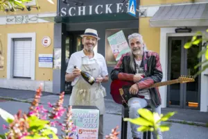 Zwei Männer stehen vor einem Geschäft. Einer trägt Schürze und Hut und hält eine Flasche sowie zwei Gläser in der Hand, der andere sitzt mit einer roten E-Gitarre auf einem Hocker. Vor ihnen steht ein Werbeaufsteller für „Summer in the City 2026“.