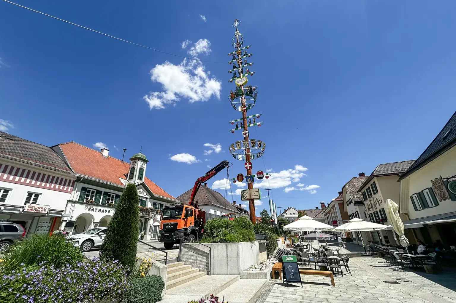 Marktplatz mit Rathaus, Gastgärten und einem hohen, geschmückten Zunftbaum. Daneben steht ein orangefarbener Lkw mit Kran. Über dem Platz spannt sich ein wolkenloser blauer Himmel.