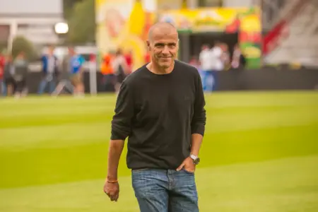 Ein Mann in schwarzem Pullover und Jeans geht über einen Fußballplatz und blickt in die Kamera. Im Hintergrund sind unscharf Zuschauer und Veranstaltungsstände am Spielfeldrand zu erkennen.
