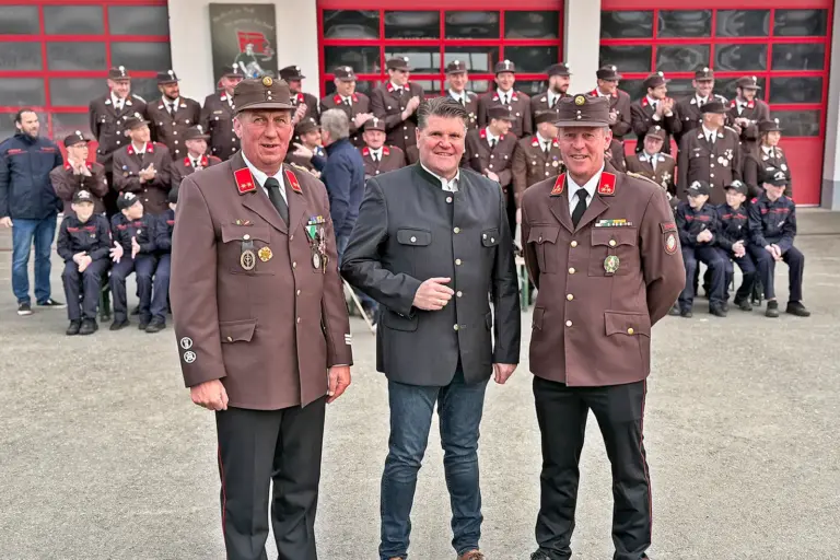 Zwei Männer in Uniform und ein Mann im Trachtensakko stehen vor einer Gruppe von Feuerwehrmännern