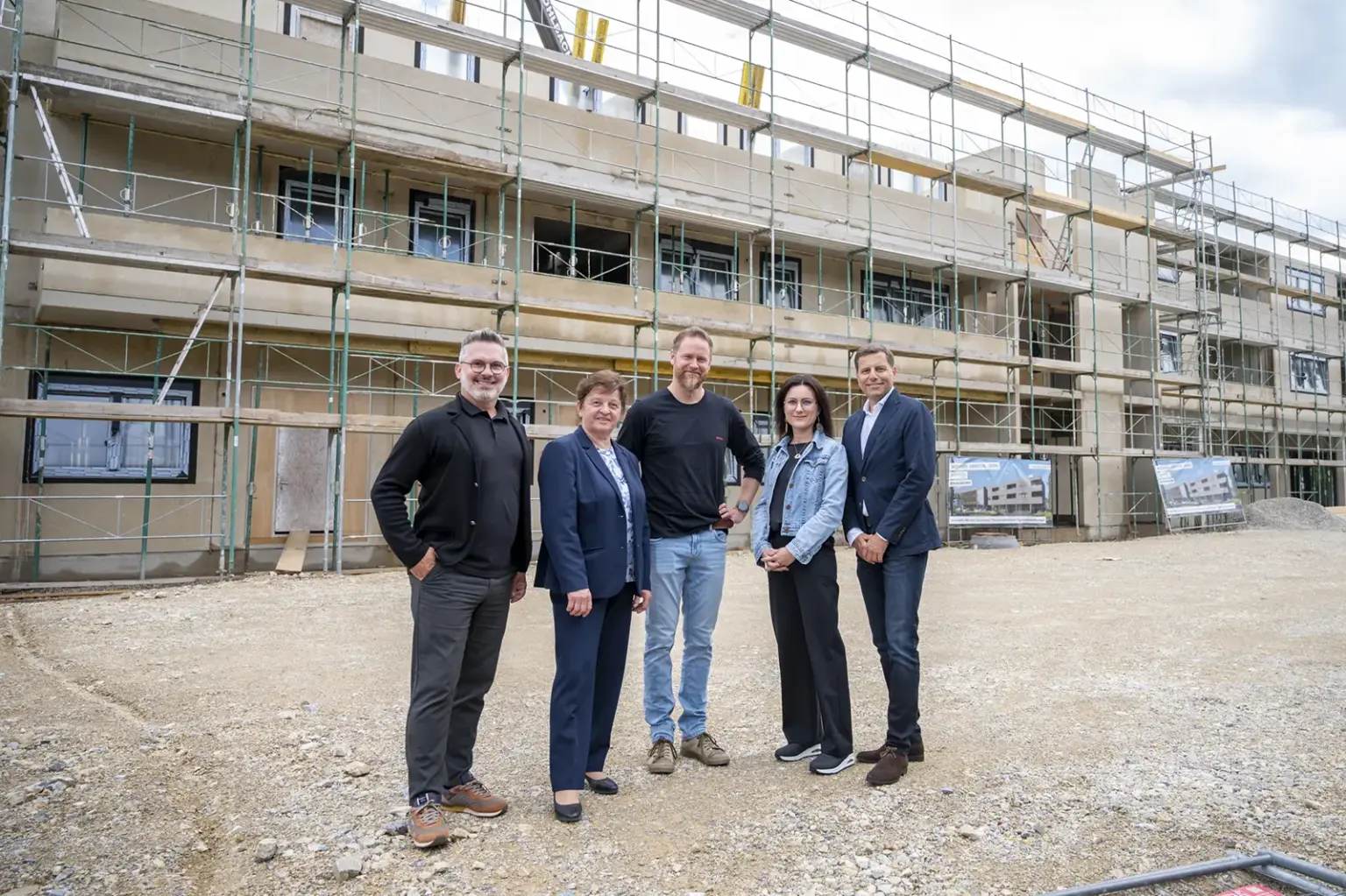 Wohnen, arbeiten und leben im Krieglacher Zentrum Vor einem mehrgeschossigen Wohnbauprojekt in Krieglach stehen fünf Personen auf dem Baustellengelände. Hinter ihnen erstreckt sich das im Bau befindliche Gebäude mit Gerüsten über die gesamte Fassade. Fenster und Balkone sind bereits erkennbar, während die Arbeiten an der Außenhülle noch andauern. Die Gruppe blickt in die Kamera, im Vordergrund liegt eine geschotterte Freifläche. Das Foto dokumentiert den Baufortschritt eines modernen Wohnprojekts im Ortszentrum.