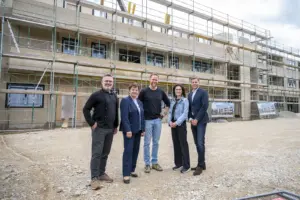 Vor einem mehrgeschossigen Wohnbauprojekt in Krieglach stehen fünf Personen auf dem Baustellengelände. Hinter ihnen erstreckt sich das im Bau befindliche Gebäude mit Gerüsten über die gesamte Fassade. Fenster und Balkone sind bereits erkennbar, während die Arbeiten an der Außenhülle noch andauern. Die Gruppe blickt in die Kamera, im Vordergrund liegt eine geschotterte Freifläche. Das Foto dokumentiert den Baufortschritt eines modernen Wohnprojekts im Ortszentrum.