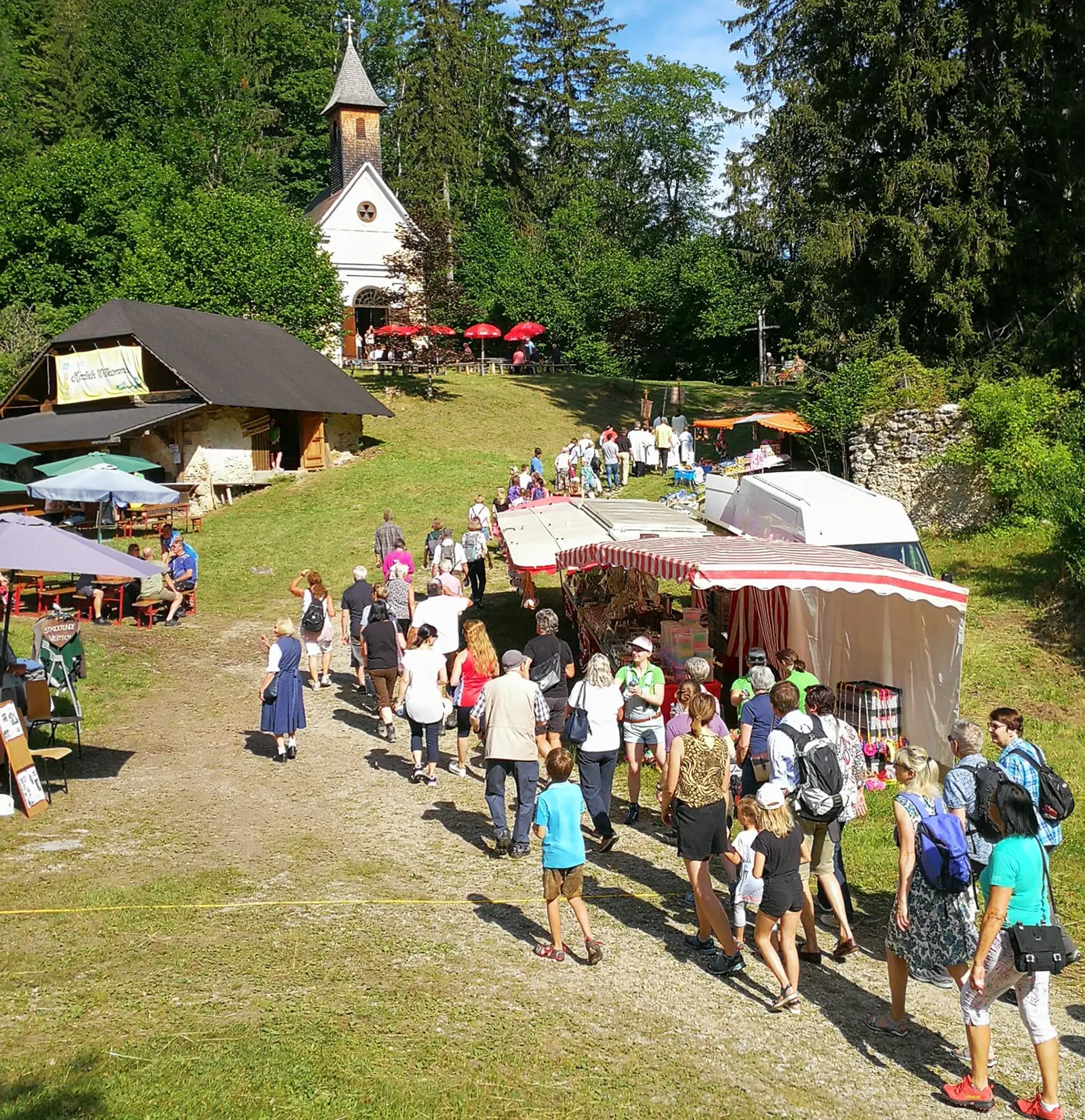 Eine Gruppe von Personen spaziert zwischen Kirtagsständen vor einer Kirche auf einem Berg