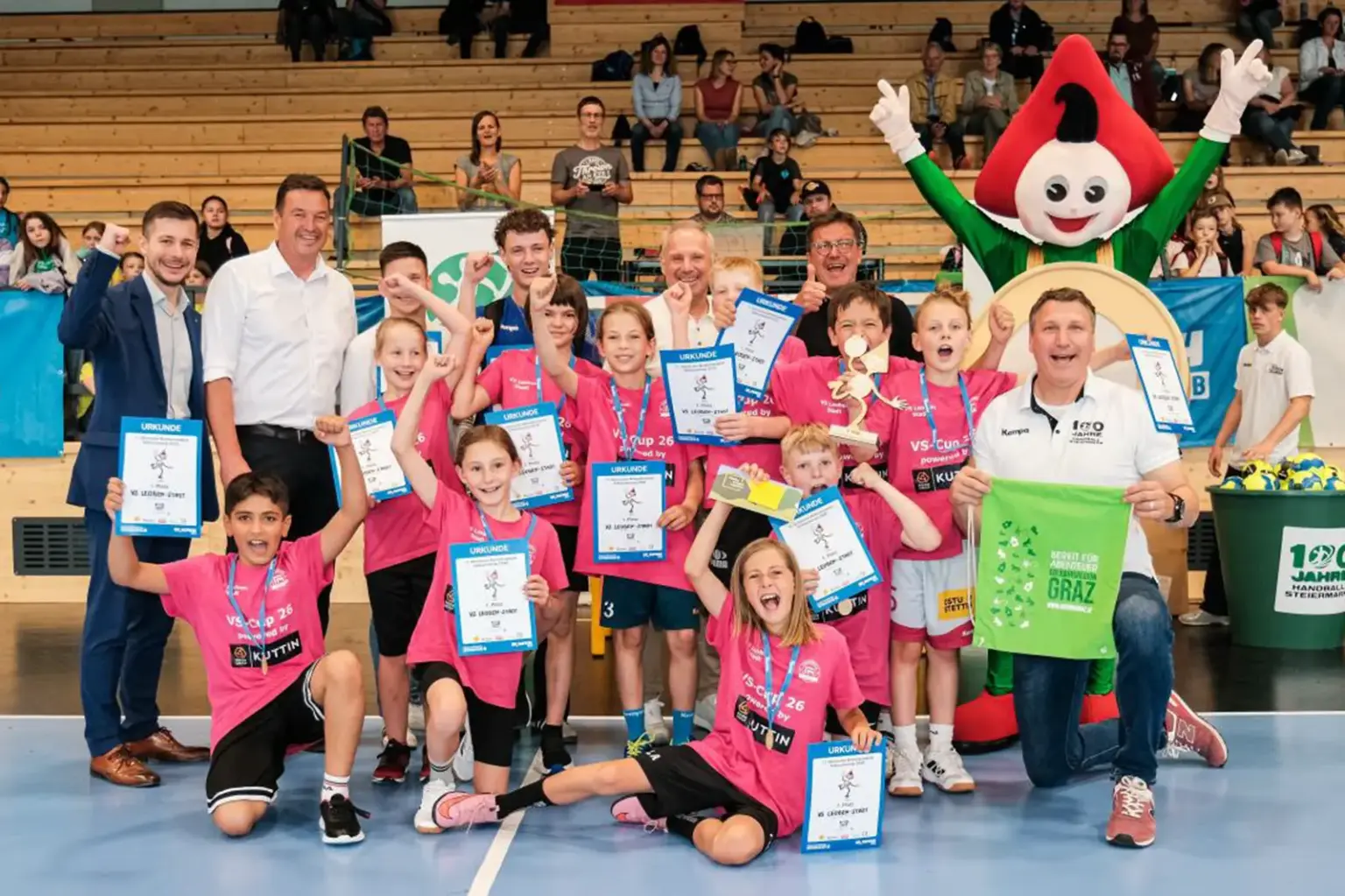 Kinder einer Handballmannschaft feiern in einer Sporthalle mit Urkunden, Medaillen und Pokal. Gemeinsam mit Betreuern und einem Maskottchen posieren sie jubelnd für ein Gruppenfoto.