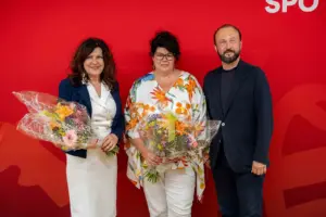 Zwei Frauen mit Blumensträußen und ein Mann stehen vor einer roten Wand