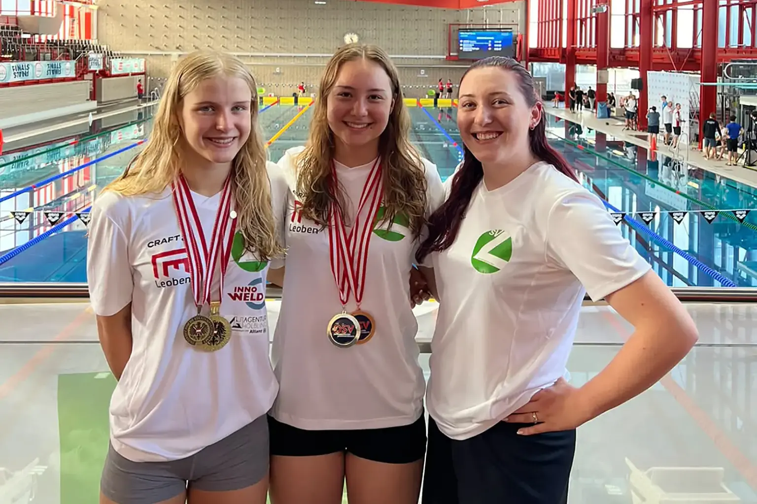 Drei Schwimmerinnen des SV Leoben präsentieren ihre Medaillen vor einem Hallenbad.