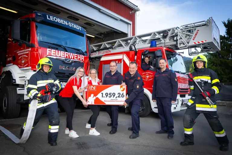 Zwei Feuerwehrleute in Einsatzbekleidung und fünf weitere Personen stehen vor zwei roten Feuerwehrfahrzeugen vor einer Fahrzeughalle. In der Mitte halten mehrere Personen gemeinsam einen symbolischen Spendenscheck, während ein weiterer Feuerwehrmann im Fahrerhaus eines Hubrettungsfahrzeugs sitzt.