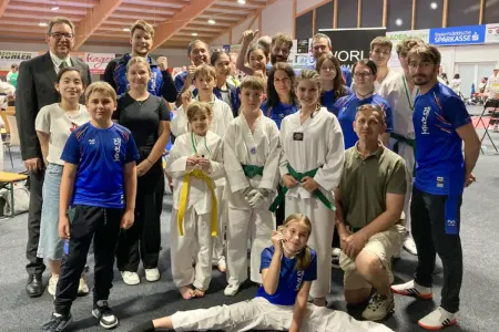 Mitglieder eines Taekwondo-Vereins präsentieren sich nach einem Turnier für ein Gruppenfoto in einer Sporthalle. Kinder, Jugendliche und Erwachsene in Trainingskleidung und Kampfanzügen zeigen Medaillen und posieren gemeinsam mit ihren Betreuern.