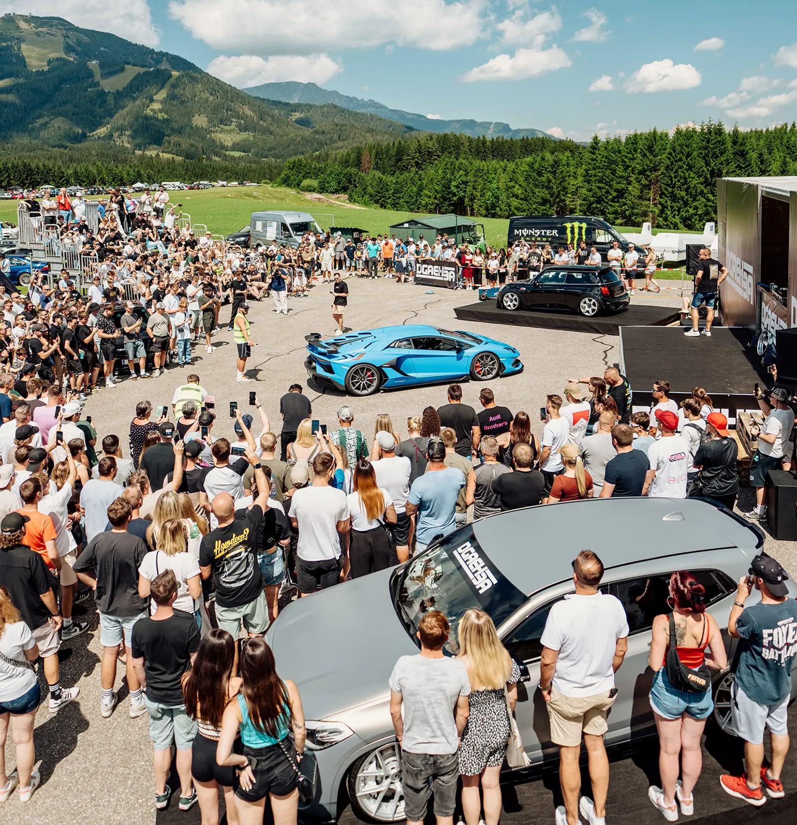 Ein großer Platz mit mehreren Autos und vielen Personen vor einem Bergpanorama