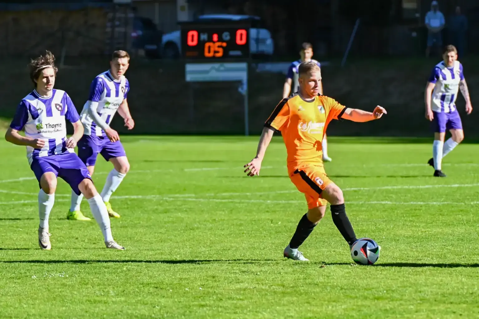 Das sind die Duelle im Steirercup Ein Fußballspieler im orangefarbenen Trikot führt den Ball über das Spielfeld, während mehrere Gegenspieler in violett-weißen Dressen seine Bewegung beobachten. Im Hintergrund zeigt die Anzeigetafel einen Spielstand von 0:0.