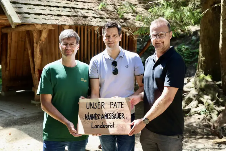Drei Männer vor einem Holzhaus präsentieren eine Holztafel