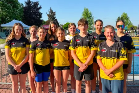 Zehn Kinder und Jugendliche in schwarz-gelben Vereinstrikots stehen für ein Gruppenfoto vor einem Freibad. Im Hintergrund sind Schwimmbecken, Absperrgitter, Zelte und Bäume zu sehen.