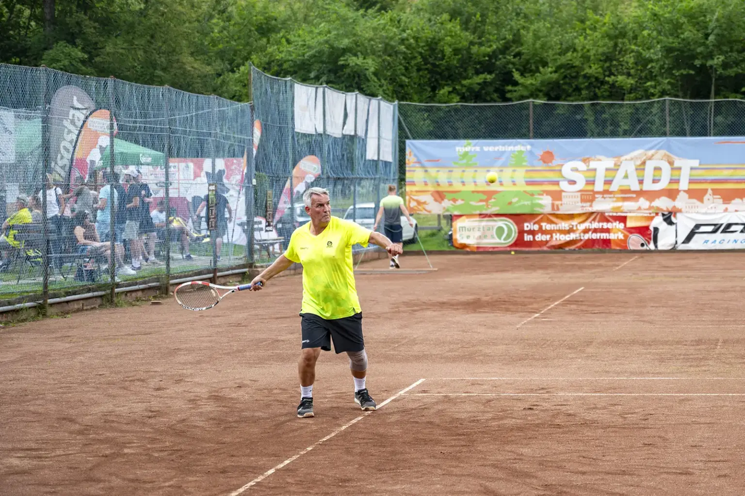 Ein Tennisspieler in einem neongelben Shirt und schwarzen Shorts schlägt auf einem Sandplatz einen Ball mit der Vorhand. Im Hintergrund sind Zuschauer hinter einem Zaun sowie Werbebanner entlang des Tennisplatzes zu sehen.