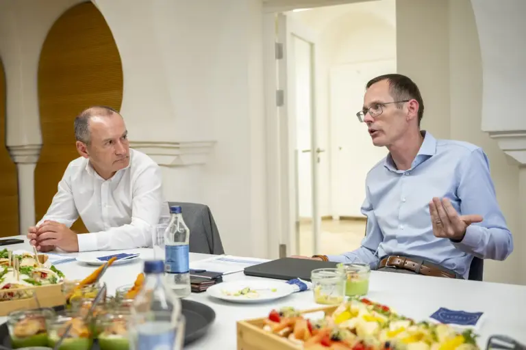 Zwei Männer sitzen an einem Besprechungstisch mit Getränken, Unterlagen und kleinen Speisen. Der Mann rechts spricht und gestikuliert, während der Mann links ihm aufmerksam zuhört.