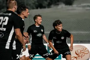 Mehrere Fußballspieler in schwarzen Dressen beobachten konzentriert eine Spielsituation auf dem Fußballplatz. Die Akteure stehen in Erwartung des nächsten Spielzugs vor einem Zaun und einer ländlichen Kulisse.
