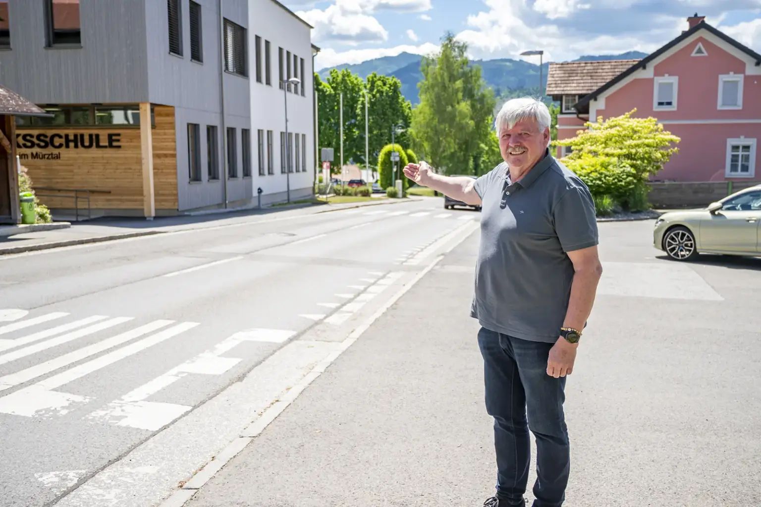 Ein Mann steht vor einer Volksschule an einer Straße und zeigt auf einen Bereich im Ortszentrum. Im Hintergrund sind Wohnhäuser, Bäume und die Berglandschaft zu sehen.