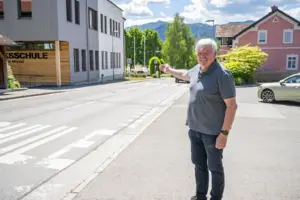 Ein Mann steht vor einer Volksschule an einer Straße und zeigt auf einen Bereich im Ortszentrum. Im Hintergrund sind Wohnhäuser, Bäume und die Berglandschaft zu sehen.
