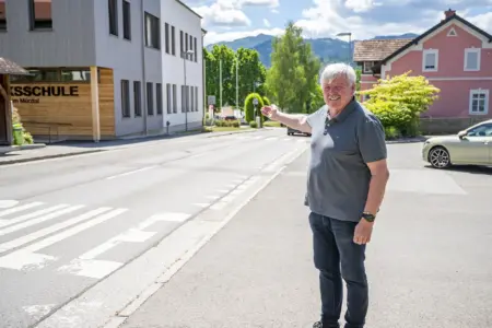 St. Lorenzen: Rad- und Gehwege sind in Planung Ein Mann steht vor einer Volksschule an einer Straße und zeigt auf einen Bereich im Ortszentrum. Im Hintergrund sind Wohnhäuser, Bäume und die Berglandschaft zu sehen.
