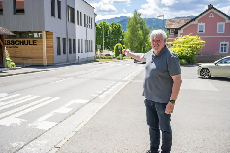 St. Lorenzen: Rad- und Gehwege sind in Planung Ein Mann steht vor einer Volksschule an einer Straße und zeigt auf einen Bereich im Ortszentrum. Im Hintergrund sind Wohnhäuser, Bäume und die Berglandschaft zu sehen.