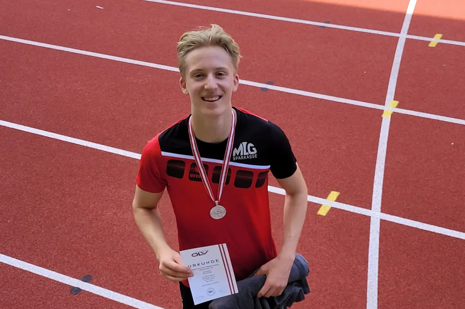 Junger Leichtathlet steht mit Medaille und Urkunde auf einer Laufbahn und blickt in die Kamera.