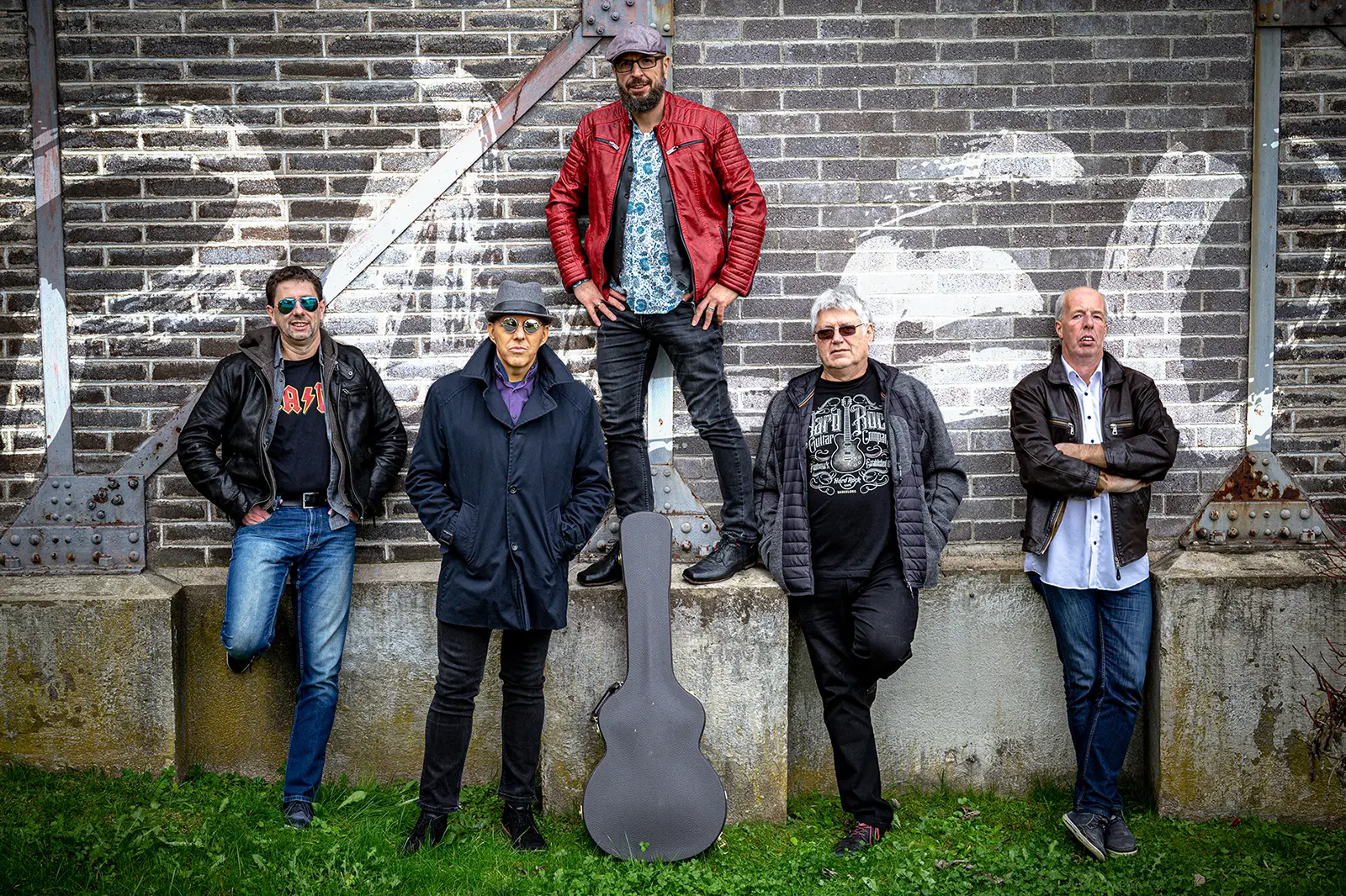 Fünf Männer stehen mit einem Gitarrenkoffer vor einer Ziegelmauer