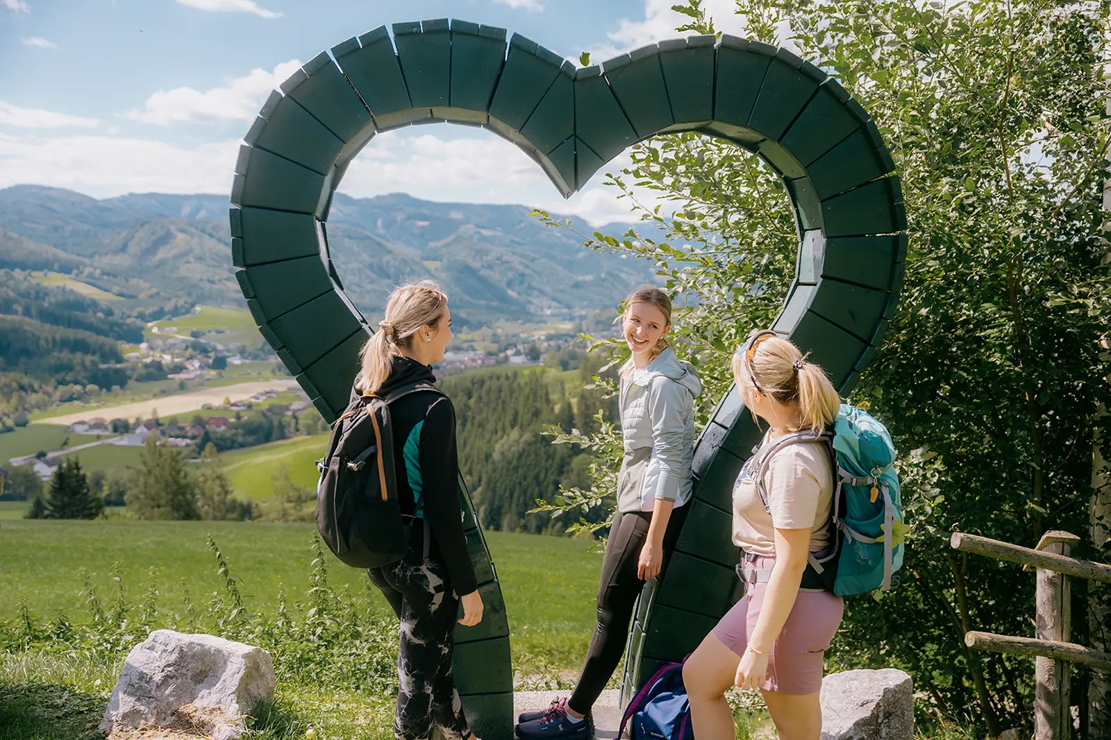 Drei Wanderinnen mit Rucksäcken stehen vor einem großen Herz in der Landschaft