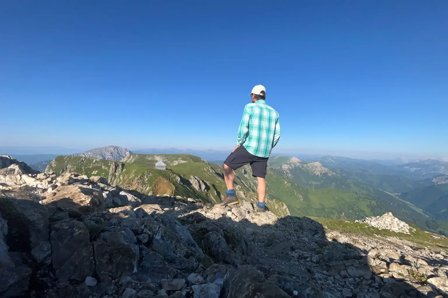 Urlaub zu Hause: Was das Rundschau-Team im Sommer treibt Eine Person mit Kappe, kariertem Hemd und kurzer Hose steht auf einem felsigen Berggipfel und blickt über eine weitläufige Gebirgslandschaft. Im Vordergrund liegen Felsen, im Hintergrund sind Bergkämme, grüne Almen und ein wolkenloser blauer Himmel zu sehen.