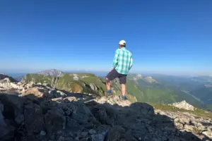 Eine Person mit Kappe, kariertem Hemd und kurzer Hose steht auf einem felsigen Berggipfel und blickt über eine weitläufige Gebirgslandschaft. Im Vordergrund liegen Felsen, im Hintergrund sind Bergkämme, grüne Almen und ein wolkenloser blauer Himmel zu sehen.