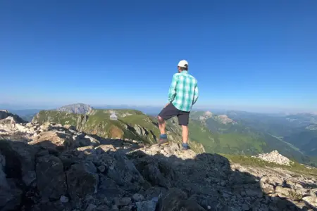 Urlaub zu Hause: Was das Rundschau-Team im Sommer treibt Eine Person mit Kappe, kariertem Hemd und kurzer Hose steht auf einem felsigen Berggipfel und blickt über eine weitläufige Gebirgslandschaft. Im Vordergrund liegen Felsen, im Hintergrund sind Bergkämme, grüne Almen und ein wolkenloser blauer Himmel zu sehen.