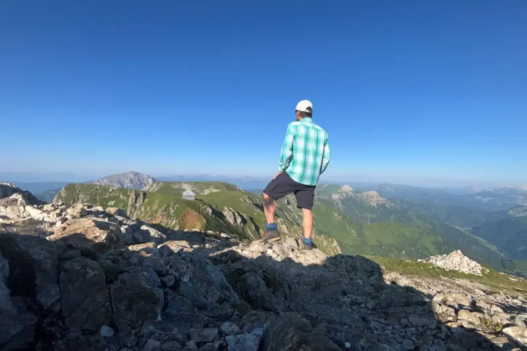 Eine Person mit Kappe, kariertem Hemd und kurzer Hose steht auf einem felsigen Berggipfel und blickt über eine weitläufige Gebirgslandschaft. Im Vordergrund liegen Felsen, im Hintergrund sind Bergkämme, grüne Almen und ein wolkenloser blauer Himmel zu sehen.