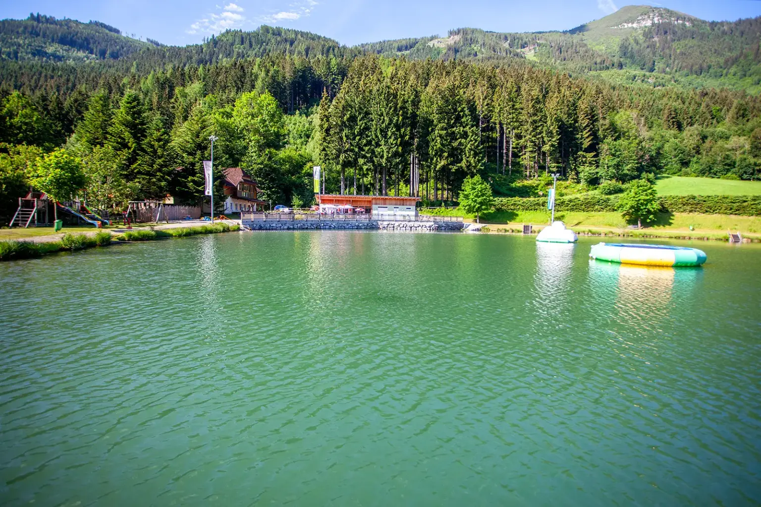 Neue Gastropächterin am Walder Freizeitsee Ein Badesee mit Trampolin- und Kletterinsel, am Ufer Liegewiese, Gebäude und ein Spielplatz, im Hintergrund Wald und Berge