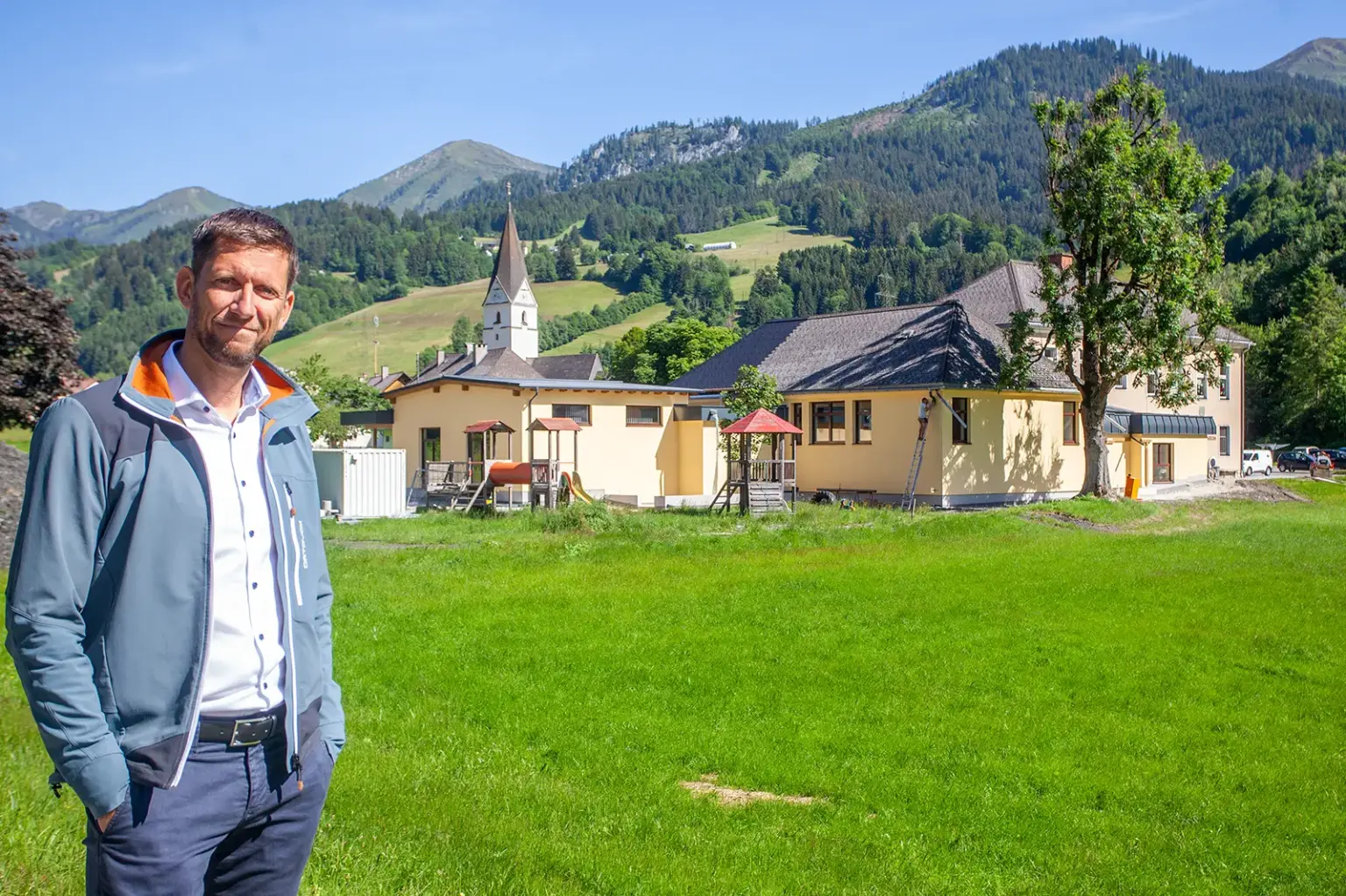 Erneuertes Haus für Kinder und Vereine in Wald am Schoberpaß Ein Mann steht auf einer Wiese vor einem Gebäude mit gelber Fassade, im Hintergrund ein Kirchturm und Berge