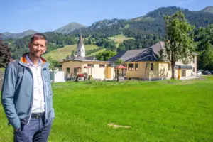 Ein Mann steht auf einer Wiese vor einem Gebäude mit gelber Fassade, im Hintergrund ein Kirchturm und Berge