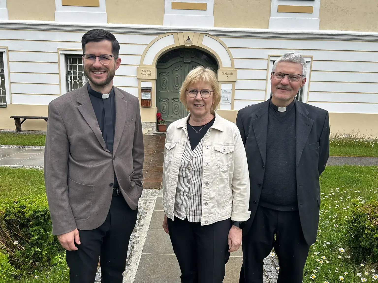 Aufschwingen zu neuen Taten Zwei Priester und eine Frau stehen vor einem hellen Gebäude