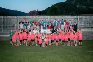 Große Gruppe von Kindern und Erwachsenen auf einem Fußballplatz bei einer Benefizveranstaltung. Im Vordergrund stehen Kinder in leuchtend pinken T-Shirts, dahinter Organisatoren und Unterstützer mit einem Spendenscheck über 5.786 Euro. Im Hintergrund sind Tribüne, Zaun und bewaldete Hügel zu sehen.