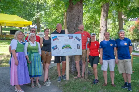 Zehn Personen stehen bei einer Veranstaltung im Freien vor einem großen Baum und präsentieren eine Infotafel zum 410-jährigen Vereinsjubiläum. Einige tragen Tracht oder Sportbekleidung, im Hintergrund sind ein Festzelt und Parkanlagen zu sehen.