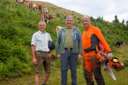 Eifrige Freiwillige halfen auf den Almen Ein Mann in Trachtenhemd und Lederhose, ein Mann in sportlicher Bergkleidung und ein Mann mit Sicherheitskleidung und Motorsäge auf einer Bergweide, im Hintergrund Kühe